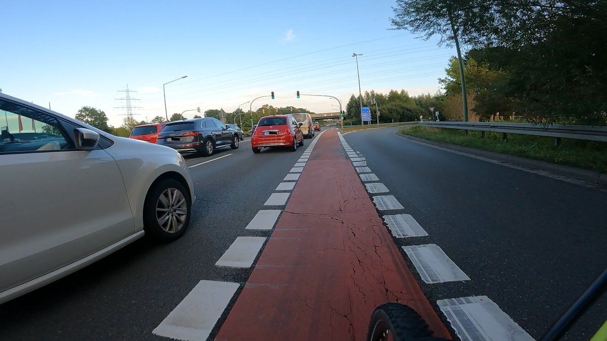 Rot markierter Radfahrstreifen zwischen einer Geradeaus-Spur mit einem KFZ welches Eng überholt. Die Ampel ist grün. Der rechte Auffahrtstreifen zur Autobahn ist noch leer.