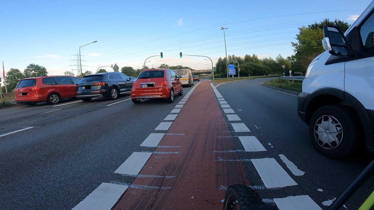 Rot markierter Radfahrstreifen zwischen einer Geradeaus-Spur mit einem roten KFZ welches gerade Eng überholt hat. Die Ampel ist grün. Auf dem rechten Auffahrtstreifen zur Autobahn überholt ein Transporter ebenfalls zu eng.